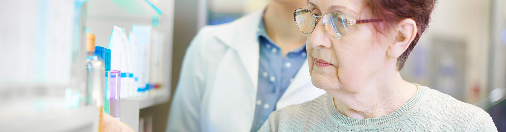 Banner featuring senior woman looking at medications at retail pharmacy with pharmacist behind her